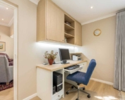 A small home office space with a desk, computer, and a blue chair, featuring shelves and a clock on the wall.