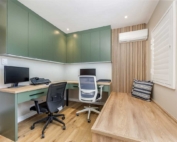 A modern home office with green cabinetry, two computer workstations, and a beige wall with wooden accents.